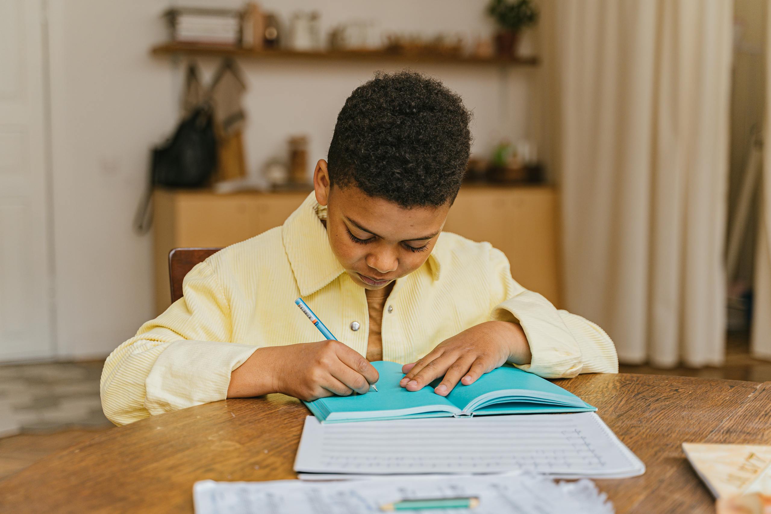A young boy focuses on writing in his notebook while seated indoors during the day.